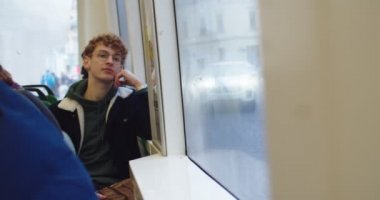 Caucasian young guy student in glasses and with red hair sitting at the tram or bus and looking at the window.