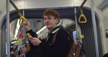 Portrait shot of the young caucasian red-haired guy student in glasses tapping on the smartphone while standing in the tram or bus, then turning face and smiling to the camera.