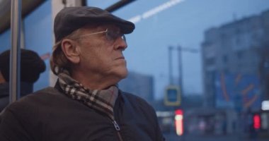 Caucasian old mn in glasses and hat sitting at te window and going home in the tram or bus in the evening. Close up.