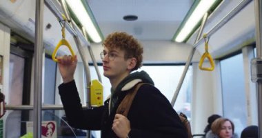 Portrait shot of the young Caucasian guy with red hair and in glasses standing in the tram and bus and looking at the camera.