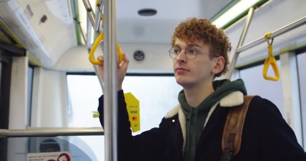 Retrato do jovem estudante caucasiano ruivo de óculos virando o rosto e olhando para a câmera enquanto vai a algum lugar no bonde ou ônibus . — Vídeo de Stock