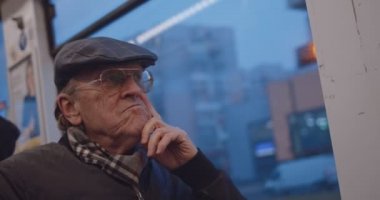 Close up of the old Caucasian man pensioner in glasses, scarf and hat going home in the evening on the tram or bus while sitting at the window and thinking.