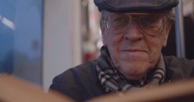 Close up of the face of the old Caucasian man in glasses and hat who smiling while reading a book in the tram or bus.