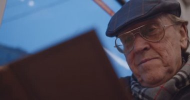 Close up of the old Caucasian man in glasses and hat reading a book while coming back home in the tram or bus in the evening.