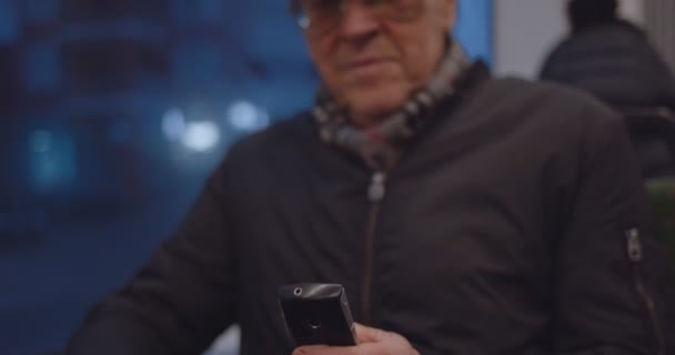 Caucasian senior man in hat and glasses taping on the old phone when sitting at the window in the evening in the tram or bus. — Stock Video