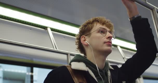 Close up do jovem estudante caucasiano cara em óculos e fones de ouvido ouvir a música e ir a algum lugar enquanto está de pé no bonde ou ônibus . — Vídeo de Stock