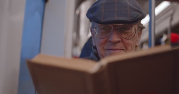 Retrato del viejo caucásico sonrió hombre guapo en gafas y sombrero sentado en la ventana por la noche en el tranvía o autobús y leyendo interesante libro . — Vídeos de Stock