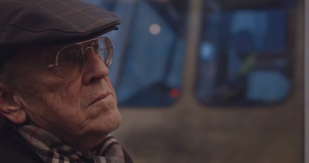 Közelkép a szemüveges, sálas és kalapos idős férfi arcáról, aki este hazafelé jövet ül a villamoson vagy a buszon.. — Stock videók