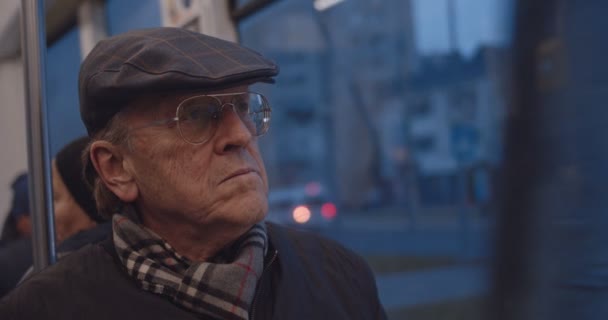 Retrato del hombre caucásico mayor guapo en gafas, bufanda y sombrero sentado en la ventana en el tranvía o autobús por la noche, luego girando la cara a la cámara con una sonrisa. De cerca. . — Vídeos de Stock