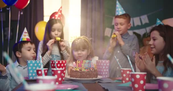 Klein schattig meisje blaast de kaarsen op haar gelukkige verjaardag taart en haar vrolijke vrienden staan rond de tafel en vieren met haar op kinderfeestje. — Stockvideo