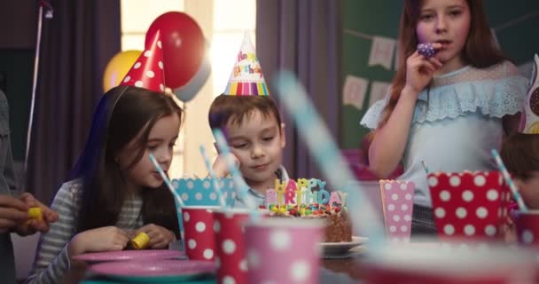 Sevimli küçük beyaz çocuk masanın üzerine eğilmiş ve doğum günü partisinde arkadaşlarının arasında pastaya bakmaktan sıkılmış.. — Stok video