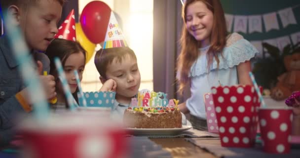 Close up of the cheerful small Caucasian boy sitting at the table with a Happy Birthday cake on and smiling while kids blowing in the party horns. — 비디오