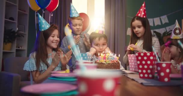 Pequeño cumpleañero caucásico soplando las velas en el pastel entre sus amigos alegres en la fiesta genial para niños . — Vídeos de Stock