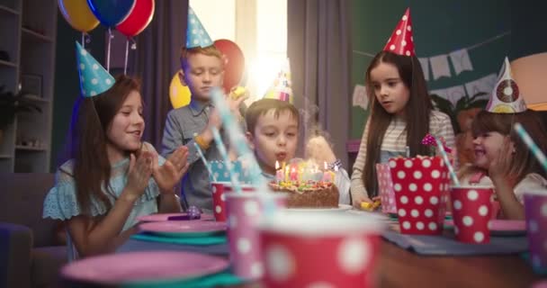 Caucásico pequeño lindo chico en el cono de cumpleaños sentado a la mesa entre los niños alegres y soplando velas en el pastel en su fiesta para amigos . — Vídeo de stock