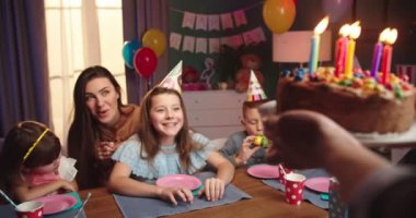 Arkadaşları ve annesiyle birlikte masada oturan ve elinde mumlarla doğum günü pastasını bekleyen beyaz bir kız..