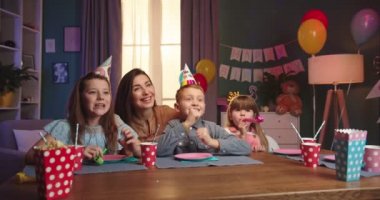 Doğum günü kutusundaki neşeli beyaz çocuk arkadaşları ve annesiyle masada oturuyor ve birisi masaya pasta getirirken bekliyor, sonra da mumları üflüyor..