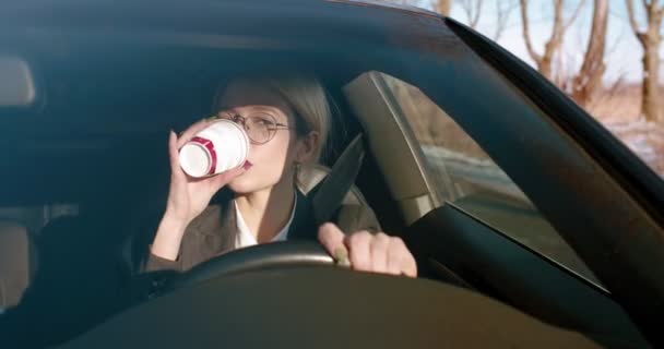 Πολύ κομψή νεαρή λευκή επιχειρηματίας με γυαλιά οδηγεί ένα αυτοκίνητο και πίνει καφέ ενώ τραγουδάει και ακούει ραδιόφωνο.. — Αρχείο Βίντεο