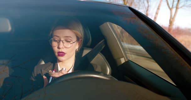 Hübsche kaukasische junge blonde Geschäftsfrau, die Lippen mit rotem Lippenstift bemalt, während sie in den Spiegel schaut und im teuren Auto sitzt. — Stockvideo