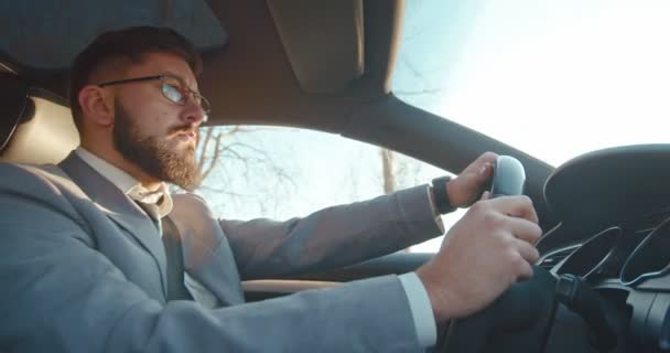 Side view on the young Caucasian serious businessman in glasses and costume driving a car and then smiling. Sunny day. — 비디오