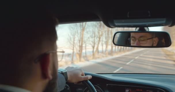 Pohled zezadu na bělocha v brýlích, jak řídí auto na silnici a dívá se do zrcadla. Zadek. Pohled přes rameno. — Stock video