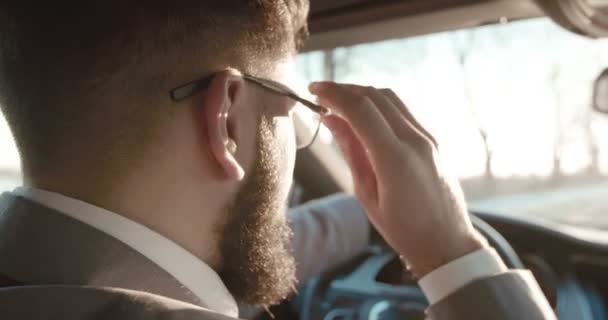 Задняя часть на белом человеке с бородой и в костюме чинит очки рукой за рулём автомобиля. Закрывай. Вид через плечо . — стоковое видео