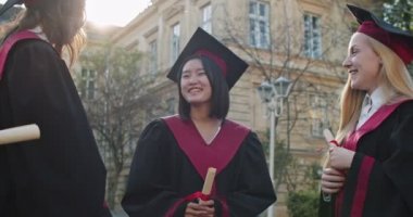 Çok ırklı, neşeli, akademik kıyafetler içinde mezun olmuş üniversite bahçesinde elinde diplomalarla neşeli bir şekilde konuşuyor, gülümsüyor ve şakalaşıyor. Açık havada..