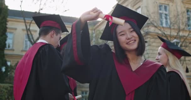 Portraitaufnahme der süßen jungen asiatischen Studentin in akademischer Mütze und Kleid, die am Abschlussabend vor der Kamera posiert, ihr Diplom in die Kamera zeigt und den Daumen nach oben streckt. — Stockvideo
