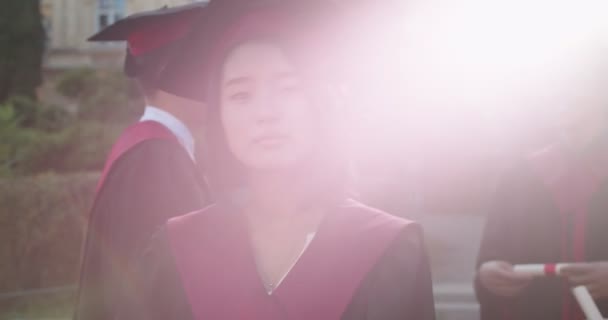 Akademik şapkası ve cübbesi olan genç Asyalı güzel kız öğrencinin mezuniyet töreninde ayakta dikilirken gün ışığında yüzünü kameraya çevirmesini yakından çek. Portarit. Açık hava.. — Stok video
