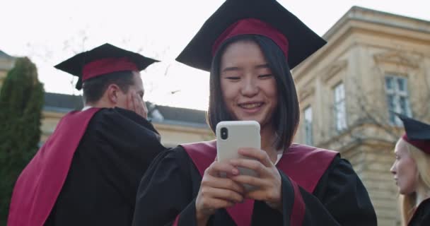 Close up da estudante asiática formada em um boné e vestido de pé ao ar livre o edifício da universidade e usando smartphone, batendo, rolagem, mensagens de texto. Recebendo parabéns — Vídeo de Stock