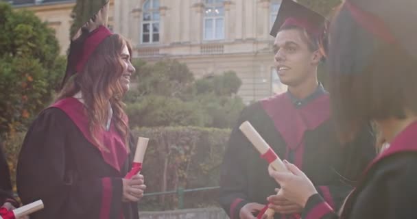 Giovani maschi e femmine di razze miste studenti gioiosi laureati in berretti e abiti con certificati in mano che parlano e ridono mentre stanno fuori dall'università dopo la cerimonia . — Video Stock