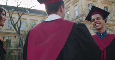 İki çok ırklı genç arkadaş, akademik kıyafetli mezun öğrenciler, kucaklaşıyorlar, gülümsüyorlar ve birbirlerini mezuniyetten sonra kutluyorlar..