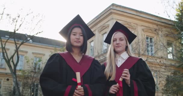 Çok ırklı genç kızların portresi, güzel üniversite öğrencilerinden akademik şapkalar ve elbiseler içinde mezun olmuş, elinde diplomalarla kameranın önünde durmuş ve gülümsüyor.. — Stok video