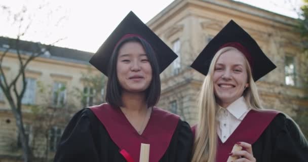 Ritratto delle giovani ragazze carine di razze miste laureate all'università in berretti accademici e abiti con diplomi in mano che posano la macchina fotografica e sorridono felicemente. Migliori amici del liceo . — Video Stock