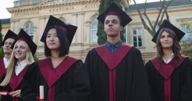 Akademik şapkalı, cüppeli, elinde diplomalarıyla mezuniyet gününde kameraya gülümseyen mutlu çoklu etnik öğrencilerin portresi..