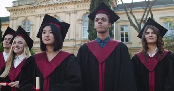 Portrait des jeunes étudiants multiethniques heureux en casquettes et robes académiques et avec des diplômes dans les mains debout à la cour de l'université et souriant à la caméra le jour de la remise des diplômes . — Video