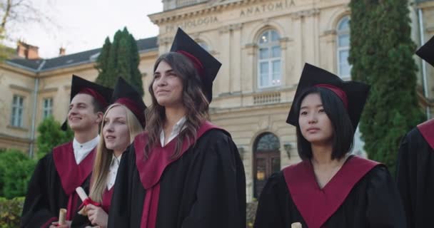 Retrato de los jóvenes y alegres estudiantes de carreras mixtas en atuendos académicos y con diplomas en las manos de pie en el patio de la universidad y sonriendo a la cámara en el día de la ceremonia de graduación . — Vídeos de Stock