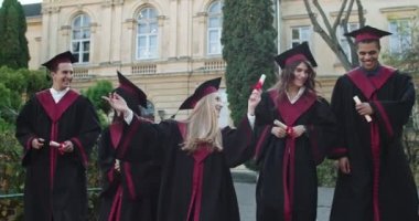 Bir grup neşeli mezun, karma ırk kızları ve akademik şapkalı ve cüppeli erkekler üniversitenin diploma ve sertifika töreninden sonra mutlu oluyorlar. Açık hava..