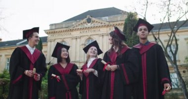 Genç, çoklu etnik çeşitliliğe sahip akademik kıyafetler içinde üniversite binasının önünde dikilen ve diploma ve resmi tören sonrası havaya şapkalar atan öğrenciler..