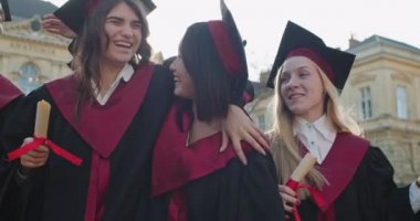 Mezuniyet töreninden sonra neşeli, çoklu etnik gruptan mezun genç kızları yakından takip edin. Çok iyi arkadaşlar, mezuniyet töreninden sonra diplomalarıyla gülüp sarılıyorlar. Açık hava..