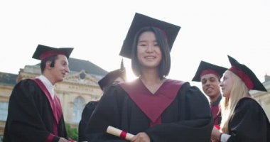 Genç ve güzel Asyalı bir kızın portresi, yüksek lisans öğrencisi akademik kıyafetler içinde ve diploması elinde kameraya gülümseyip şapkasını havaya fırlatıyor. Açık hava..