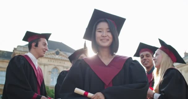 Portret van het jonge mooie Aziatische meisje, master student afgestudeerd in de academische outfit en met diploma in handen glimlachend naar de camera en dan gooien in de lucht haar pet. Buiten. — Stockvideo