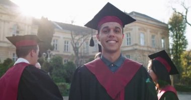 Genç ve yakışıklı bir adamın portresi, yüksek lisans öğrencisi akademik şapkalı ve cüppeli üniversite binasında kameraya neşeyle gülümseyen. Açık havada. Kapat..