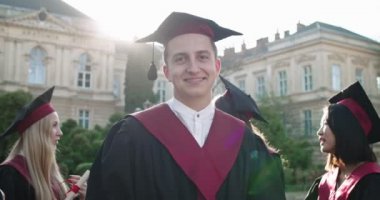 Genç ve neşeli beyaz adamın portresi, akademik şapkalı ve cüppeli mezun öğrenci, üniversite binasındaki kamerada da gülümsüyor. Açık havada. Kapat..