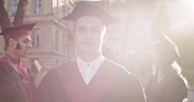 Akademik şapkası ve cübbesi olan, üniversitenin önünde kameraya gülümseyen komik, genç, beyaz, mutlu erkek öğrenciyi yakından çek. Portre.
