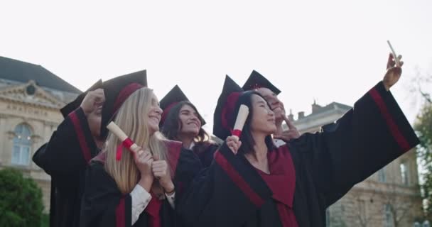 Grupo de jóvenes mestizos graduados en gorras académicas y vestidos con diplomas en las manos sonriendo alegremente a la cámara mientras toman fotos de selfies en el teléfono inteligente. Exterior . — Vídeo de stock