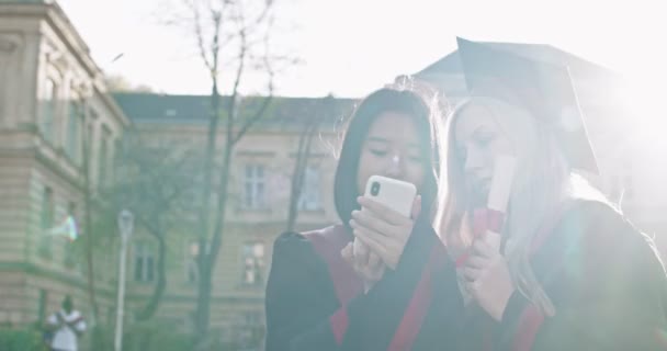 Close up of the two multiethnic young best friends girls, pretty master graduates with diplomas and in academic caps. One girl showing to another photos, chat or video on the smartphone. Outdoors. — Stock Video