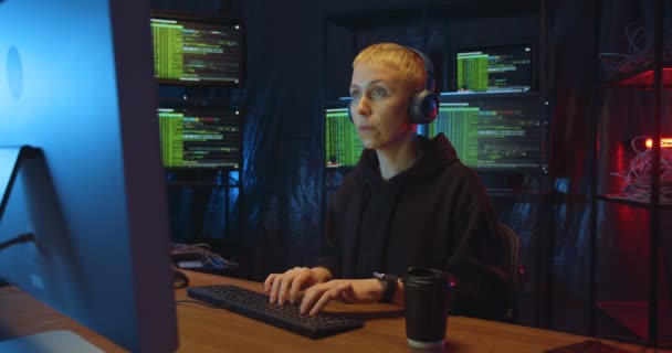 Hacker mujer caucásica en los auriculares que trabajan en la computadora y escribir en el cuarto oscuro. Mujer bebiendo café mientras comete delitos cibernéticos. Beber bebida caliente mientras está en línea por la noche . — Vídeo de stock