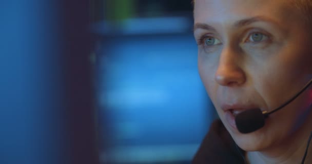 Close-up van een blanke vrouw van middelbare leeftijd in een headset met microfoon terwijl ze belt met een cliënt in een donkere kamer met monitoren. Vrouwelijke werknemer van call-center voor computerscherm. — Stockvideo