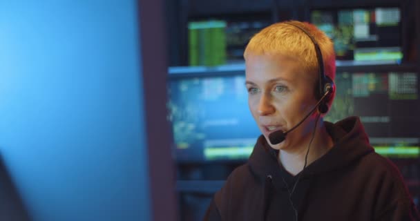 Biała kobieta w średnim wieku z krótkimi blond włosami i słuchawkami mówiącymi w mikrofonie podczas pracy na ekranie komputera w centrum monitoringu połączeń w ciemnym pokoju w nocy. Kobieta policjantka ds. bezpieczeństwa cybernetycznego. — Wideo stockowe