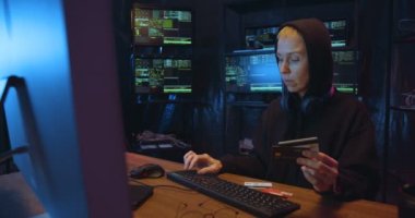 Kapüşonlu beyaz kadın hacker karanlık bilgisayar odasında kredi kartlarıyla dolandırıcılık yaparken finansal siber suç işliyor. Klavyeye numara ve parola yazan kadın girişi.
